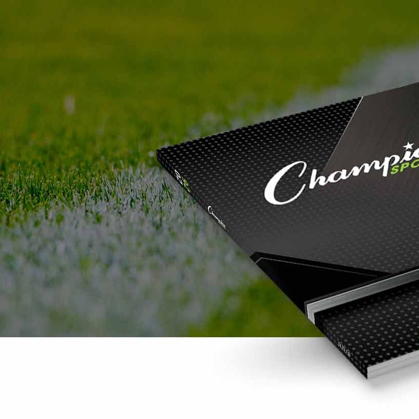 A black sports magazine or book titled 'Champion Sports 2026' with green accents, shown resting on a blurred green grass field with a white line.